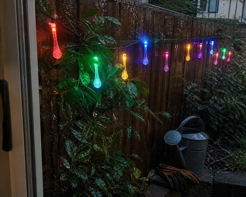 Multicolor teardrop lights glowing along a wet garden fence after rain